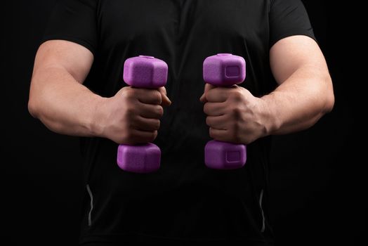 adult male athlete in black uniform holds blue plastic purple dumbbells in hands, black background