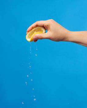 female hand holds half a yellow lemon and squeezes it on a blue background, splashes fly to the sides