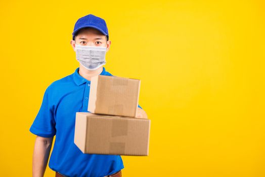 Front of Asian young delivery worker man in blue t-shirt and cap uniform wearing face mask protective giving cardboard boxes under coronavirus or COVID-19, studio shot isolated yellow background
