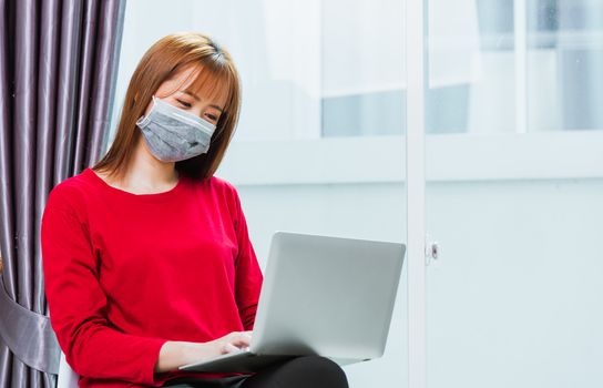 Asian young business woman wear red t-shirt sitting on chair wearing face mask protective working from home office using laptop computer he quarantines disease coronavirus or COVID-19 with copy space