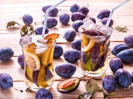 Refreshing drink from fresh juicy sweet plums with lemon in glass mugs with ice cubes on a wooden background with plums and green leaves