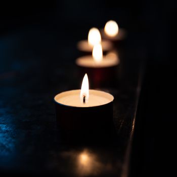 Tea candles in a church resting on a dark surface reflect their light, square color image