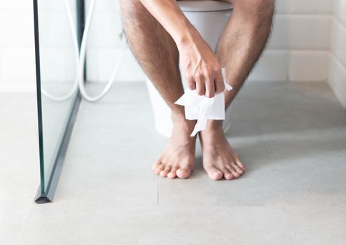 Closeup man sitting on toilet with tissue paper in hand, selective focus