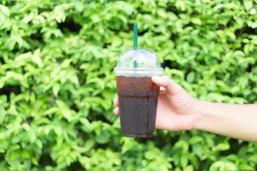 Close up hand holding glass of ice americano coffee with green leaves nature background, selective focus