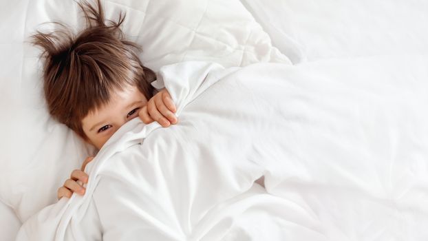 Cute toddler hides in bed. Disheveled boy wakes up and peeps out from under white blanket. Morning bedtime at cozy home.