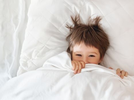 Cute toddler hides in bed. Disheveled boy wakes up and peeps out from under white blanket. Morning bedtime at cozy home.