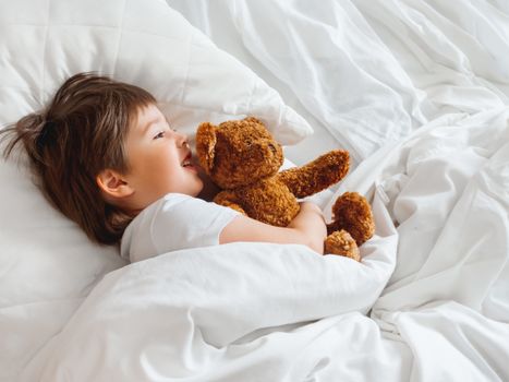 Toddler lies in bed with cute teddy bear. Little boy under white blanket with fluffy toy. Plush guard watches out child's sleep. Morning bedtime at cozy home.