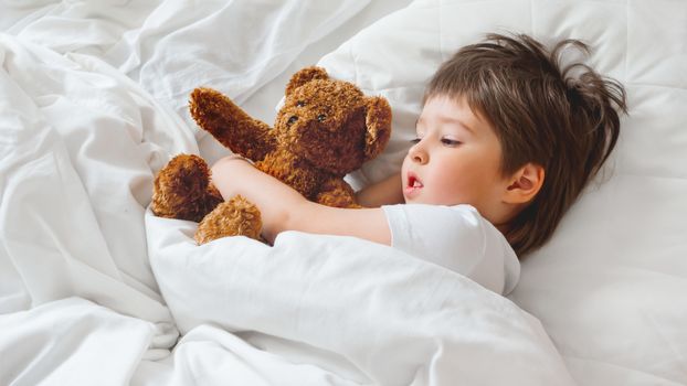 Toddler lies in bed with cute teddy bear. Little boy under white blanket with fluffy toy. Plush guard watches out child's sleep. Morning bedtime at cozy home.