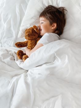 Toddler lies in bed with cute teddy bear. Little boy under white blanket with fluffy toy. Plush guard watches out child's sleep. Morning bedtime at cozy home.