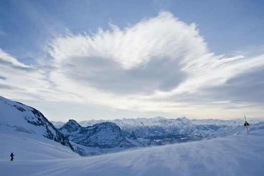 A heart shaped cloud floats over a ski run as part of an expansive mountain vista in the italian alps