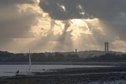 A man pushes his yacht up a jetty on a stony beach, with factory industrial towers behind him in southern wales
