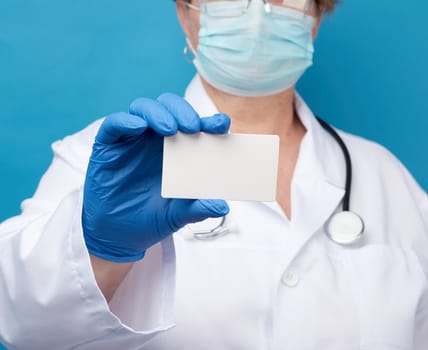 doctor hand in blue medical latex gloves holds an empty paper business card on a blue background, place for an inscription