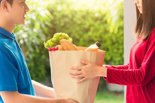 Asian young delivery man in uniform making grocery fast service giving fresh food in paper bag to woman customer receiving at house door under pandemic coronavirus, Back to new normal concept