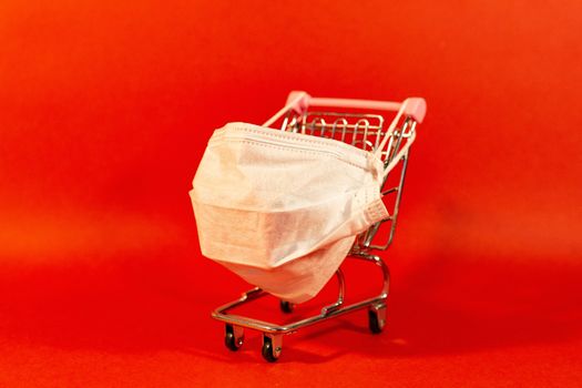 Shopping cart with protective medical mask against coronavirus. Safe and online shopping on quarantine concept. Red background.