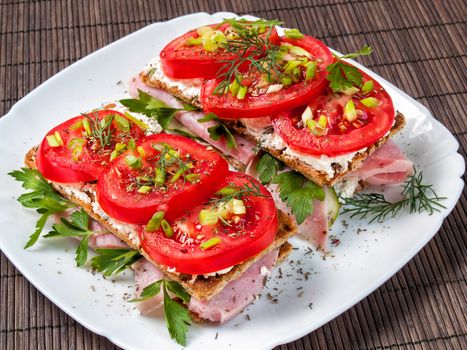 On a white plate close-up lie sandwiches from crunchy buckwheat loaves smeared with curd cheese on top is a tomato, ham, greens, spices