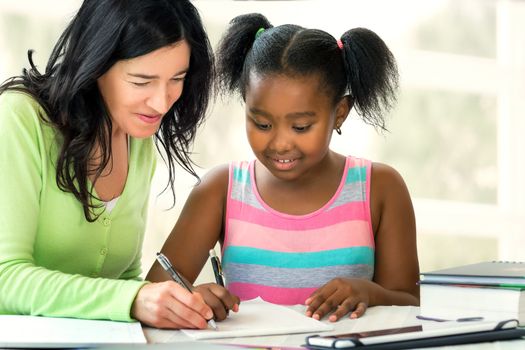 Close up portrait of caucasian teacher and little african female student resolving mathematic home work. Two women writing at desk at home.