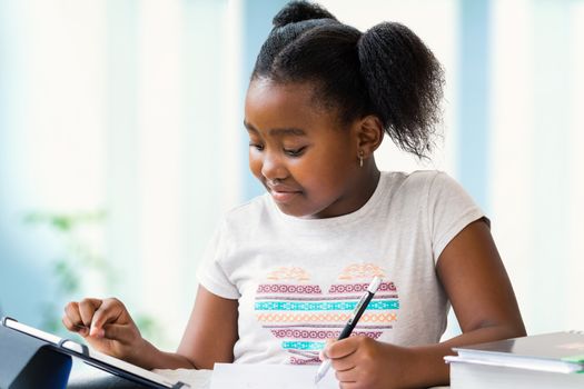Close up portrait of cute little african girl doing home work at desk. Ponytailed kid typing on digital tablet and writing with pen on paper.