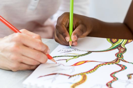 Detail of caucasian teacher drawing together with african little girl.Two hands creating sketch with color pencils.