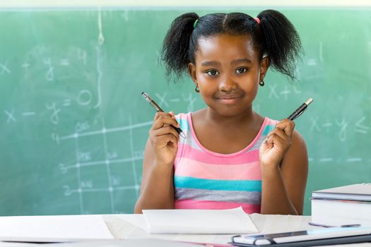 Close up portrait of young african student holding pen and pencil at desk in classroom with black board in background.