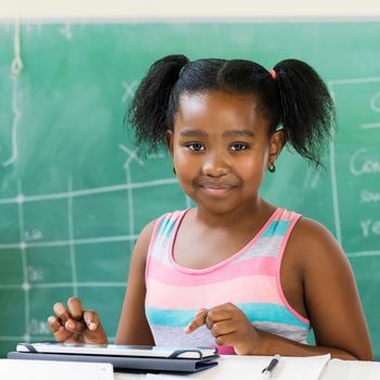 Close up portrait of little african student sitting at desk with digital tablet in classroom.Kid looking at camera against black board in background.