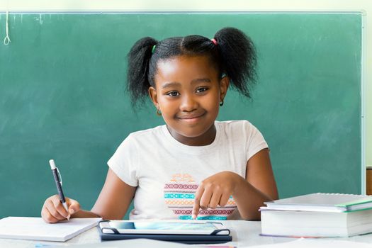 Close up portrait of cute little african student writing and working at desk with digital tablet. Ponytailed kid smiling with blank black board in background.