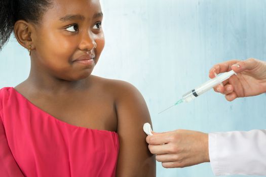 Close up portrait of anxious little african girl receiving disease prevention vaccine.Kid with nervous facial expression looking at doctor.Caucasian hands holding syringe with needle next to upper arm.