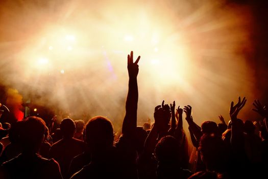 Silhouettes of festive crowd of young people dancing at music festival. Raising hands against smoky orange background with light beams.