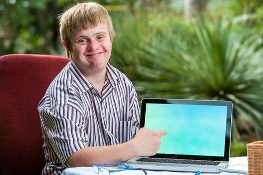 Close up portrait of friendly young student with down syndrome pointing at blank laptop screen.Sitting at desk in garden.