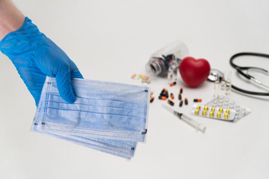 Hand holds a medical mask against a background of a red toy heart and pills. Coronavirus Protection Concept