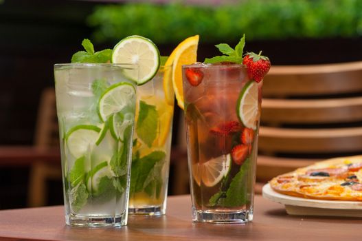 three cold drinks with citrus and strawberries in a glass on the table and pizza