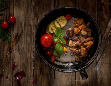 fried pieces of meat and vegetables on an old frying pan, view from above. wooden background