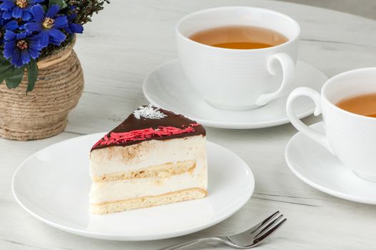 cake on a plate and two cups of tea on the table with flowers and a fork