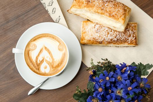 bun on paper wrapper and a cup of coffee with the spoon on a brown table with a flower