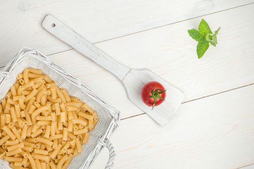 pasta in wicker basket and tomato on a wooden spoon on a white old wooden boards