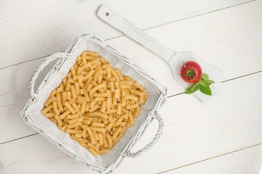 pasta in wicker basket and tomato on a wooden spoon on a white old wooden boards