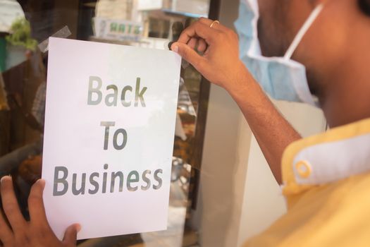 Young man with medical mask pasting Back to business poster or signage in front of store door - concept of reopening business after coronavirus or covid-19 pandemic lock down crisis