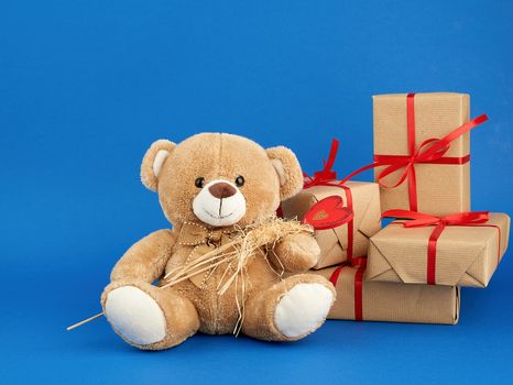 small beige teddy bear and a stack of boxes wrapped in brown kraft paper and tied with a red ribbon, blue festive backdrop for Valentine's Day and birthday