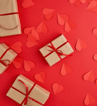 boxes wrapped in brown kraft paper and tied with red thin silk ribbon on a red background, decorative hearts are scattered nearby, holiday is Valentine's Day