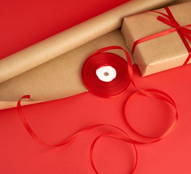 roll of brown paper for packaging, a reel of red ribbon and a wrapped gift in brown kraft paper on a red background, making surprises, place for an inscription