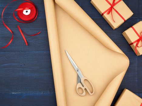 roll of brown kraft packaging paper, box tied with a red silk ribbon, scissors and a bobbin with ribbon on a blue wooden background, top view