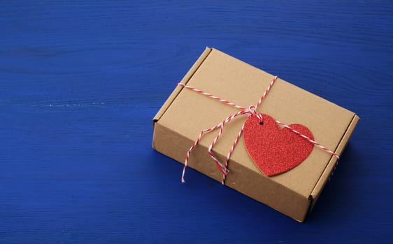 rectangular brown box with a gift and a red paper heart tied to a rope, blue wooden background, top view, copy space