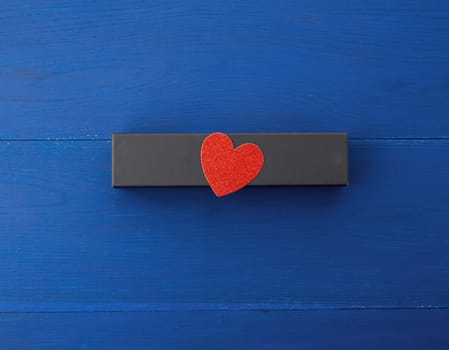 closed rectangular cardboard box with a gift, red paper heart from above, blue wooden background from boards, top view