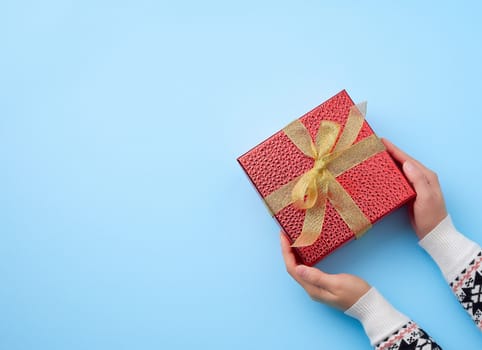 female hand holds a red box tied with a golden ribbon, concept of congratulations and gifts, top view, blue background