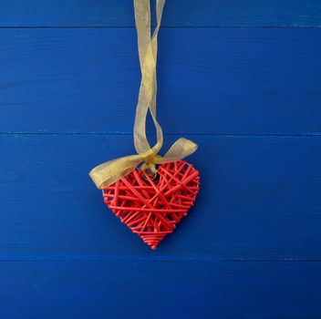 red wicker decorative heart hanging on silk golden ribbon, blue wooden festive background for Valentine's day