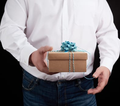man in a white shirt holds in his hand a closed brown gift box on a dark background, concept proposal of marriage, congratulations on the holiday, copy space