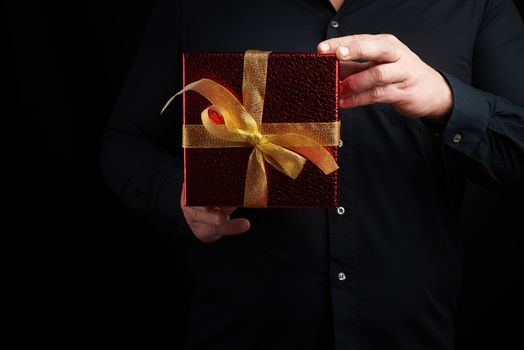 adult man in a black shirt holds a red square box with a knotted golden bow on a dark background, concept of congratulations, surprise