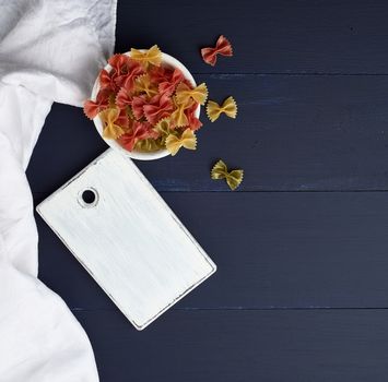 raw pasta in the form of bows in a wooden round plate and white textile napkin on a blue wooden background, top view. Mediterranean food set.