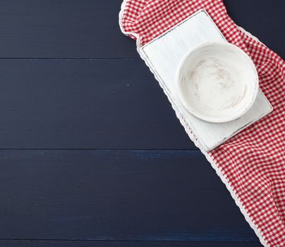 rectangular white cutting kitchen board and empty round plate on a blue wooden background, next to a white red textile towel, top view. Copy space