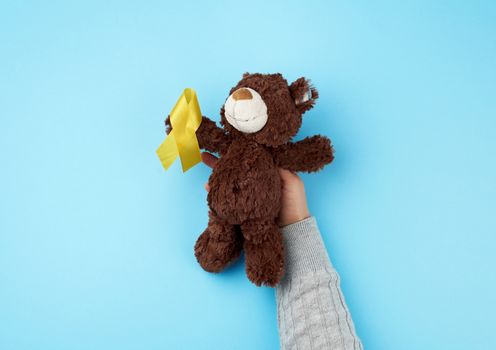 female hand hold a small brown teddy bear which holds in its paw a yellow ribbon folded in a loop on a blue background. concept of the fight against childhood cancer. problem of suicides