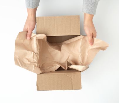 open empty square brown cardboard box for transportation and packaging of goods and two female hands lay kraft paper on the bottom, view from the top